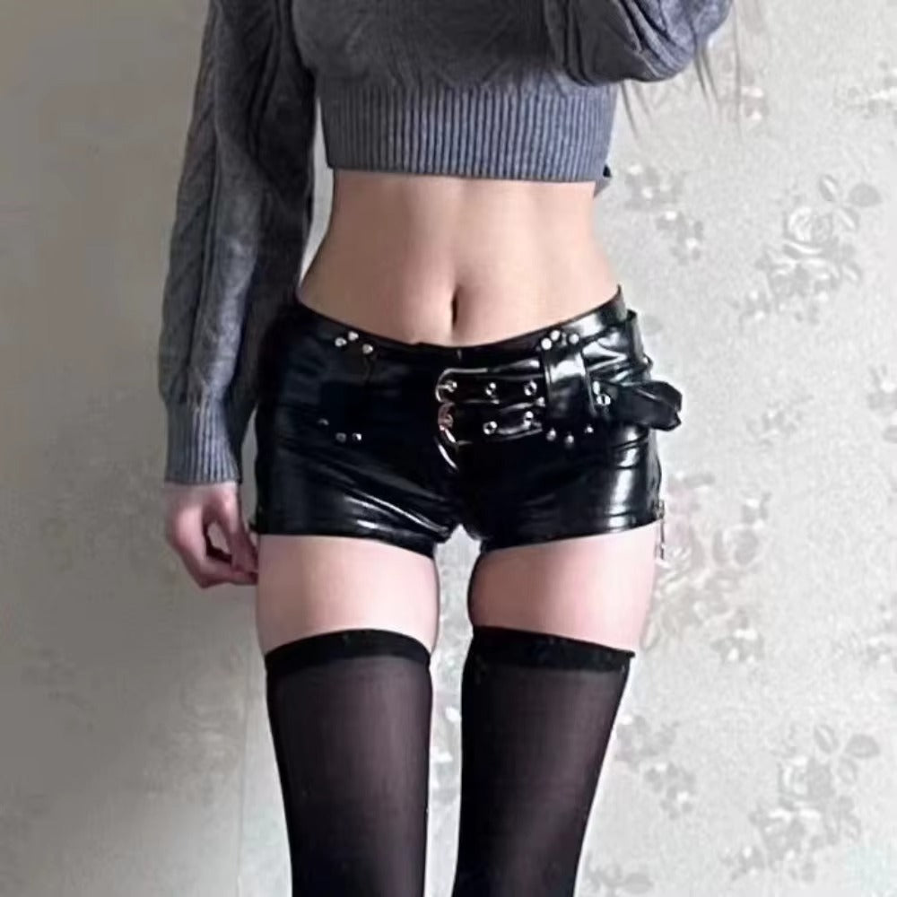 A person wearing black glossy faux-leather mini shorts with a large buckle detail, black thigh-high stockings, and a gray ribbed crop top, standing against a light patterned wall.