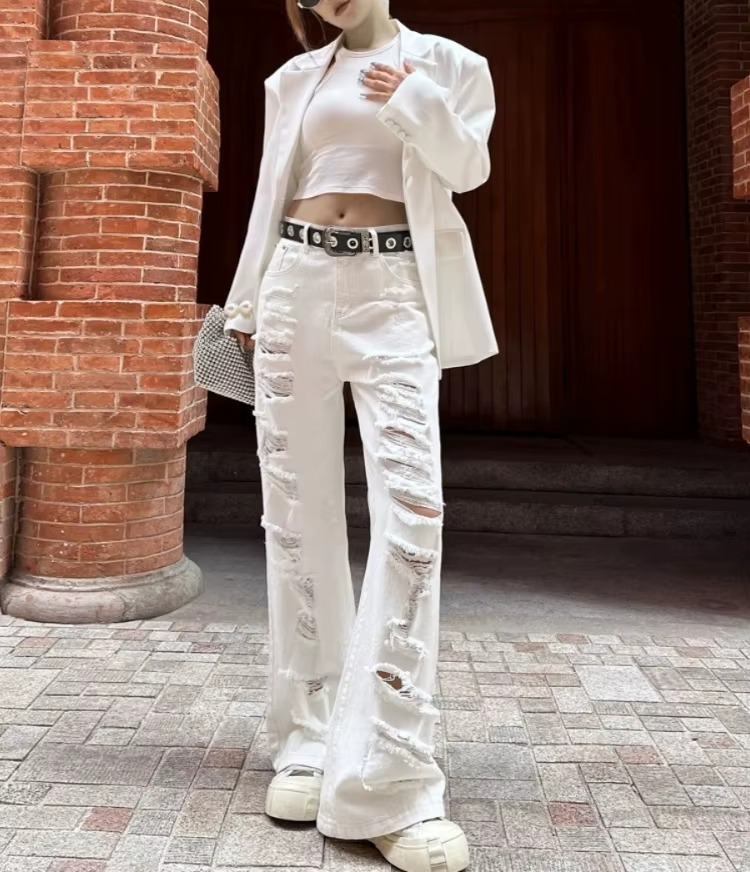 Person wearing a white outfit with ripped jeans standing against a brick wall.